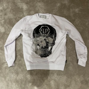 Vit sweatshirt med dödskalle från Philipp Plein - Säljer en cool vit sweatshirt från Philipp Plein med en stor dödskalle i silverpaljetter på framsidan. Tröjan har långa ärmar och en rund halsringning. Perfekt för dig som vill sticka ut med en unik design! Fick i julklapp men är tyvärr för stor. Passar dig som är lite bred👍 Värd 17500kr. Äkta har kvitto