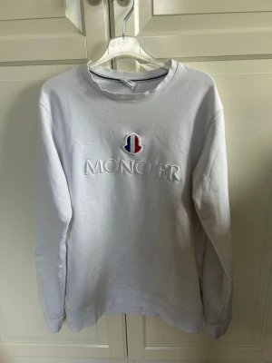 Vit sweatshirt från Moncler - Säljer en vit sweatshirt från Moncler med broderad logga i rött, blått och vitt på bröstet samt stor Moncler-text. Tröjan har rund halsringning och långa ärmar. Perfekt för dig som gillar stilrena och exklusiva plagg. Hör av dig om du har frågor tar gärna emot byten!