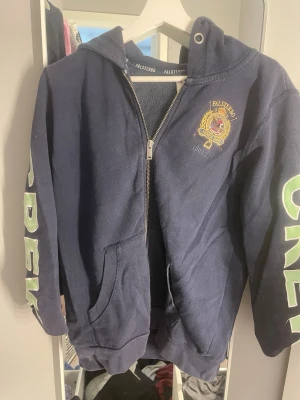 Mörkblå Falsterbo hoodie med dragkedja - Mörkblå hoodie från Gina Tricot med dragkedja, huva och fickor. På bröstet finns ett broderat Falsterbo Horse Show-emblem i gult och rött. Stora gröna bokstäver på ärmarna. Perfekt för dig som gillar hästsport och vill ha en sportig look.