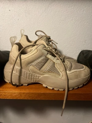 Beige chunky sneakers med meshdetaljer - Chunky sneakers i beige med meshdetaljer på sidorna och framtill. Skorna har grov sula, snörning och dragflikar bak. Materialmix av syntet och textil ger en sportig vibe. Perfekta för dig som gillar streetwear och neutrala färger.