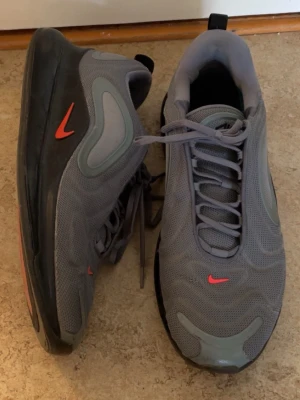 Nike Air Max 720 grå sneakers - Snygga Nike Air Max 720 sneakers i grått mesh med svarta och grå detaljer samt röda swoosh-loggor. Skorna har en chunky sula och futuristisk design med rundade former och synlig Air Max-dämpning. Perfekta för dig som gillar streetwear och sportig stil.