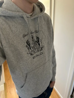 Bvaldi hoodie - Snygg grå hoodie från Bvaldi med svart tryck framtill som visar ett motiv och texten 'Côte d'Azur Club'. Hoodien har huva med dragsko och en stor magficka.    Hör av er vid frågor eller funderingar.