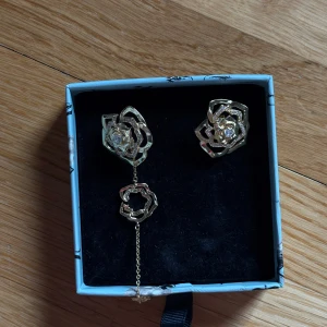 Guldiga rosformade örhängen från IOAKU - Säljer ett par unika örhängen i guldfärgad metall med rosformad design. Ena örhänget har en kedja med en extra liten ros som hänger ner. Båda har glittrande stenar i mitten som ger en lyxig känsla. Perfekt för dig som gillar detaljerade och trendiga smycken. 