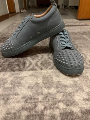 Gråa Christian Louboutin sneakers - Unika grå sneakers från Christian Louboutin med matta nitar över tån och sidan. Skorna har en låg profil, rund tå och är gjorda i mocka med matchande grå snörning. Insidan är beige och märkt med Louboutin-loggan. Perfekta för dig som vill sticka ut. Har fått dom från en släkting han hade använt som några gånger före och man ser de på bild fyra, annars bra skick. De kommer med låda. Öppen för frågor 