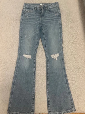 Blå utsvängda jeans från Gina Tricot - Snygga blå jeans från Gina Tricot med utsvängda ben och slitna detaljer på knäna. Jeansen har klassisk femficksmodell och råa kanter nertill. Perfekta för en trendig och avslappnad look.