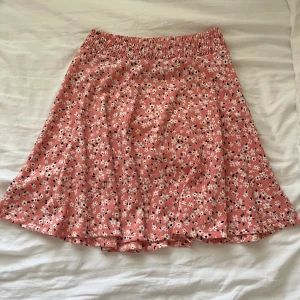 Rosa blommig kjol - Söt rosa kjol med vita och svarta småblommor och bred resår i midjan. Kjolen har en lätt utställd form och är gjord i ett mjukt, tunt tyg som ger ett flowy intryck. Perfekt för dig som gillar en romantisk och avslappnad stil. Aldrig använd. 