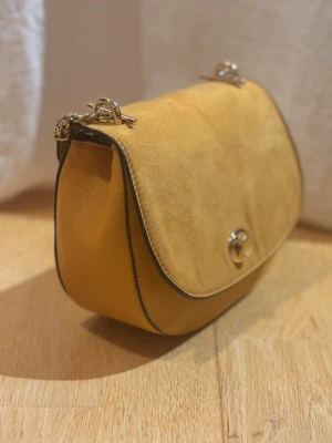 Gul väska med guld kedja  - Säljer en snygg gul axelväska i guldfärgad  kedjerem och vridlås framtill. Väskan har rundad form och stilrena detaljer, perfekt för dig som vill ha något som sticker ut. Väskan har lite fläckar på insidan tyvärr, men det är inget som syns utåt.