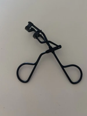 Svart ögonfransböjare i metall - En stilren svart ögonfransböjare med klassisk design och greppvänliga handtag. Tillverkad i metall med en mjuk silikonkudde för att skydda fransarna vid användning. Perfekt för att skapa snygg böj på fransarna snabbt och enkelt.