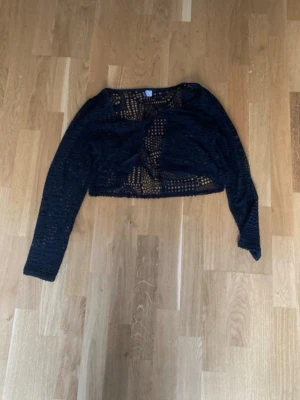 Svart virkad cropped tröja - Svart stickad cropped tröja med lång ärm och genombrutet virkat mönster. Tröjan har en avslappnad passform och är perfekt att styla över ett linne eller topp. Materialet är mjukt och luftigt, vilket gör den bekväm att bära. Det finns ett hål som är större än det andra hålen (se bild 3)