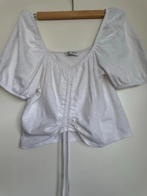 Vit omlottopp med puffärm och snörning - Trendig vit topp från Gina Tricot med puffiga korta ärmar, omlottkänsla och snörning framtill som ger en snygg rynkeffekt. Tillverkad i ett lätt, strukturerat tyg som känns luftigt och fräscht. Perfekt för dig som gillar en feminin och modern stil.