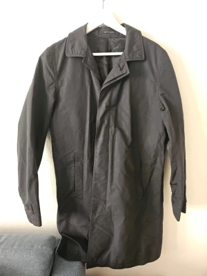 Svart trenchcoat från Tiger of Sweden - Stilren svart trenchcoat från Limonta med klassisk krage och raka linjer. Jackan har två sidofickor och är tillverkad i ett slätt tyg med italienska material. Perfekt för dig som gillar minimalistisk och tidlös design. Fattas en knapp som kan fås gratis i butik.