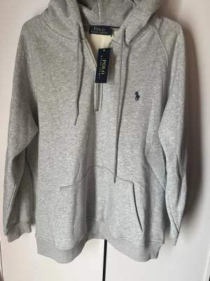 Grå hoodie från Polo Ralph Lauren - Snygg ljusgrå hoodie från Polo Ralph Lauren med halv dragkedja, huva med dragsko och klassisk blå logga broderad på bröstet. Mjuk bomullsblandning och stor känguruficka framtill. Perfekt för en chill och stilren look.