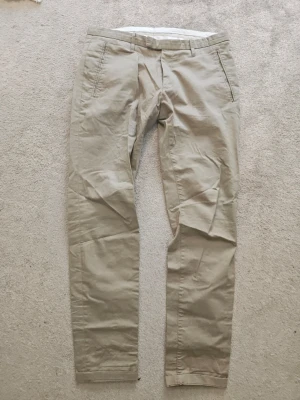 Beige chinos från Tiger of Sweden - Snygga beige chinos från Tiger of Sweden i modellen Liston 3 GW. Byxorna är tillverkade i 98% bomull och 2% elastan för en skön och stretchig passform. Klassisk rak modell med fickor i sidorna och hällor för bälte.