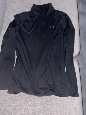 Svart träningströja från Under Armour - Snygg svart långärmad träningströja från Under Armour med halv dragkedja och hög krage. Tillverkad i ett mjukt och stretchigt funktionsmaterial som andas, perfekt för löpning eller gym. Diskret logga på bröstet.