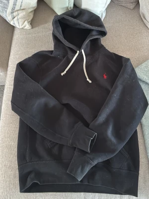 Svart hoodie från Ralph Lauren - Snygg svart hoodie från Ralph Lauren med klassisk huva och vita dragsnören. Ikoniska röda loggan broderad på bröstet. Mjuk bomullskänsla och känguruficka framtill. Perfekt för en avslappnad och stilren look.