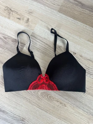 Svart bh utan bygel med röd spetsdetalj - Snygg svart bh med vadderade kupor och justerbara axelband. Framtill finns en dekorativ röd spetsdetalj med rosett som ger en extra touch. Bh:n har slät yta och är tillverkad i ett mjukt material som känns bekvämt mot huden.