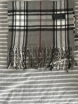 Grå rutig halsduk från Burberrys - Klassisk halsduk i grått med svart, vitt och rött rutmönster från Burberrys. Halsduken har tvinnade fransar i ändarna. Perfekt accessoar för att lyfta din outfit.