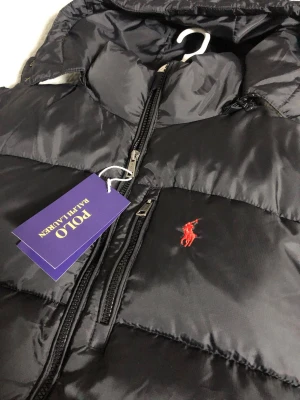 Svart pufferjacka från Polo Ralph Lauren - Snygg svart pufferjacka från Polo Ralph Lauren med huva och dragkedja framtill. Jackan har två framfickor med dragkedja och den klassiska röda loggan broderad på bröstet. Perfekt för kalla vinterdagar och riktigt stilren look.