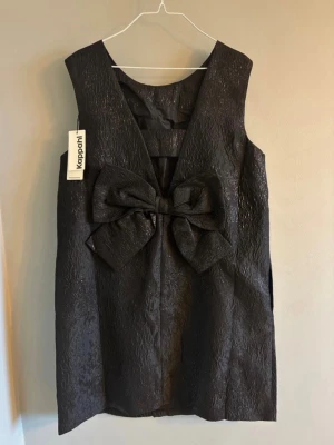 Svart kort klänning med rosett Kappahl - Snygg svart kort klänning från Kappahl i glansigt, strukturerat material. Ärmlös modell med rund halsringning och stor dekorativ rosett framtill. Perfekt för dig som vill sticka ut med en unik detalj.