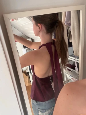 Vinröd satintopp med knyt i nacken från Gina - Säljer en vinröd topp med snygg knytning i nacken och öppen rygg. Toppen är ärmlös och har en elegant, lös passform. Perfekt att styla med jeans eller kjol för en trendig look.