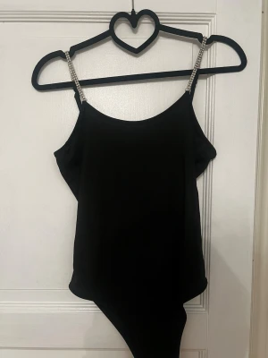 Svart body med glittriga axelband - Snygg svart body med smala axelband dekorerade med glittrande stenar. Kroppsnära passform och rundad halsringning. Materialet känns mjukt och stretchigt. 