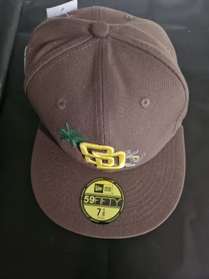 Brun New Era 59FIFTY San Diego keps - Brun keps från New Era, modell 59FIFTY, med San Diego Padres logga i gult framtill och broderad palm. Platt skärm och detaljerad brodyr på sidan. Materialet är kraftig bomull och kepsen har klassisk passform.