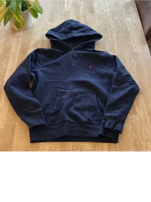 Mörkblå hoodie från Ralph Lauren - Mörkblå hoodie från Ralph Lauren med klassisk huva och magficka. Liten röd broderad logga på bröstet. Mjuk bomullskänsla och ribbade muddar vid ärmslut och nederkant. Perfekt för en avslappnad och stilren look.