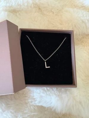 Silverhalsband med bokstav L EDBLAD - Snyggt silverfärgat halsband från EDBLAD med ett L-format hänge dekorerat med små pärlor. Kedjan är tunn och elegant, och halsbandet levereras i original ask. Perfekt för dig som vill ha en personlig touch på din stil. Skriv för frågor och bilder💕