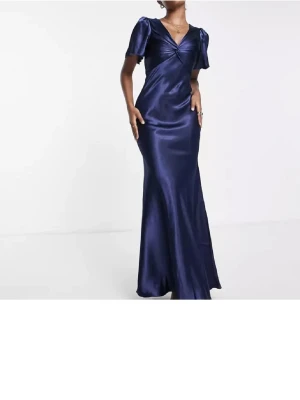 Marinblå långklänning i satin - Säljer en elegant marinblå långklänning i glansigt satinmaterial. Klänningen har korta ärmar, v-ringning och en snygg twistdetalj framtill. Perfekt för speciella tillfällen när du vill känna dig extra fin.