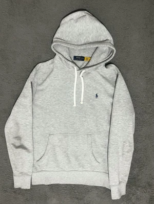Grå hoodie från Polo Ralph Lauren - Snygg ljusgrå hoodie från Polo Ralph Lauren med klassisk huva och vita dragsnören. Har en känguruficka framtill och den ikoniska lilla mörkblå loggan broderad på bröstet. Tillverkad i mjukt bomullsmaterial som känns skönt mot huden.