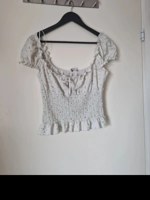 Vit blommig blus med volang - Supersöt blus med små blommor, volangkant och smockad midja. Toppen har korta puffärmar, knyt fram och en romantisk vibe. Perfekt till jeans eller kjol för en söt look. I fint skick. Köpt på Vinted men jag har endast provat den. Storlek M. Katt finns i hemmet. Jag har fler kläder och böcker till salu
