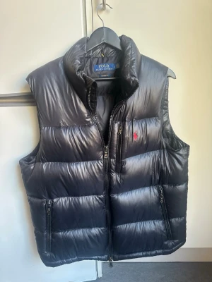 Svart dunväst från Polo Ralph Lauren - Snygg svart dunväst från Polo Ralph Lauren med hög krage och dragkedja framtill. Västen har två fickor med dragkedja och en bröstficka med röd logga. Perfekt för lager-på-lager och håller dig varm med sin vadderade design.