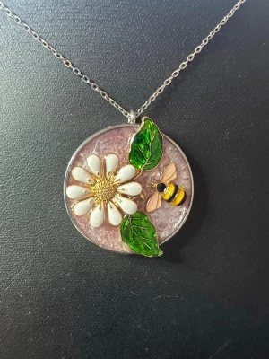 Halsband med blomma och bi - Unikt halsband med runt hänge i silverfärg, dekorerat med en vit blomma, gröna blad och en söt gul-svart bi. Bakgrunden är skimrande ljuslila. Perfekt accessoar för dig som gillar naturinspirerade smycken och detaljer.