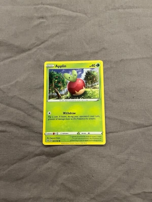 Pokémonkort Applin - Säljer ett Pokémonkort med Applin-motivet. Kortet har en grön och gul ram, med en tecknad bild av Applin i ett fruktträd. Text och symboler i svart och vitt. Perfekt för samlare eller Pokémonfans.