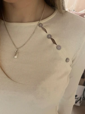 Ribbad topp med knappar Zara - Trendig ribbad topp i beige med lång ärm och unika knapprad-detaljer längs axeln. Snygg och stilren modell som passar perfekt till jeans eller kjol. Materialet känns mjukt och följsamt mot huden.