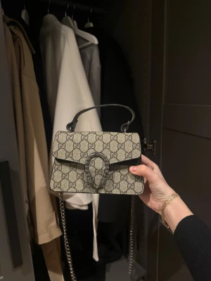  beige handväska - Säljer en beige  handväska med klassiskt  silverfärgad kedja. Väskan har ett dekorativt spänne framtill och ett kort handtag upptill. läderdetaljer och snygg rektangulär form.