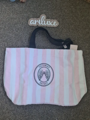 Randig shoppingväska Victoria's Secret - Säljer en stor shoppingväska från Victoria's Secret med rosa och vita ränder. Väskan har svarta handtag och ett tryckt emblem med änglavingar och logga på framsidan. Materialet känns mjukt och handduksliknande, perfekt för stranden eller shoppingrundan.helt ny och oanvänd