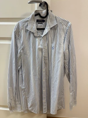 Randig skjorta från Polo Ralph Lauren - Klassisk vit skjorta med blåa ränder från Polo Ralph Lauren. Skjortan har lång ärm, knappar framtill och broderad logga på bröstet. Perfekt för en stilren look med tidlös känsla.
