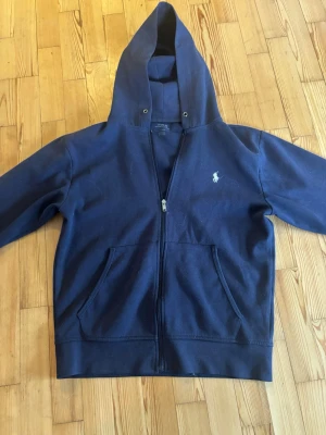 Marinblå hoodie från Polo Ralph Lauren - Snygg marinblå hoodie från Polo Ralph Lauren med dragkedja framtill och klassisk vit logga på bröstet. Hoodien har två stora fickor fram och justerbar huva. Tillverkad i mjukt bomullsmaterial som är skönt att bära.