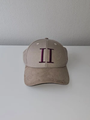 Beige keps från Les Deux - Snygg beige keps från Tommy Hilfiger med broderat vinrött 'II' framtill och små broderade detaljer i blått, vitt och rött på sidan. Klassisk böjd skärm och justerbar passform. Perfekt accessoar för en avslappnad och trendig stil.