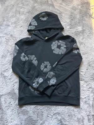 Mörkgrå hoodie med blommönster - Snygg mörkgrå hoodie med stora, ljusgrå blommönster över hela tröjan. Klassisk huva och magficka framtill. Mjuk och skön bomullsblandning, perfekt för en avslappnad streetwear-look. Ribbade muddar vid ärmslut och nederkant.