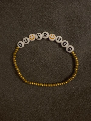 Pärlarmband med text DU ÄR NOG - Unikt armband med guldfärgade små pärlor och vita bokstavspärlor som stavar 'DU ÄR NOG', där vissa bokstäver ersatts av smileys. Perfekt accessoar för dig som vill bära ett peppande budskap på handleden.