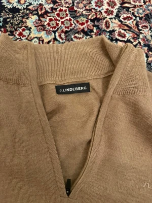 Beige stickad tröja J.Lindeberg - Snygg beige stickad tröja från J.Lindeberg med halv dragkedja och diskret broderad logga på bröstet. Tröjan har ribbade muddar och är tillverkad i mjukt material som passar perfekt till lager på lager.