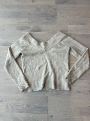 Beige off shoulder stickad tröja - Säljer en beige stickad tröja med bred off shoulder-krage och långa ärmar. Tröjan har ribbade muddar vid ärmslut och nederkant. Perfekt för dig som gillar en avslappnad men ändå trendig stil. Materialet känns mjukt och skönt mot huden.