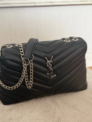 Svart YSL quiltad handväska - Säljer en svart quiltad handväska med silverfärgad kedjerem. Väskan är tillverkad i mjukt äkta läder och har ett elegant, tidlöst uttryck med chevronmönster. Perfekt för dig som gillar lyxiga accessoarer. Väldigt bra skick och kvalité.