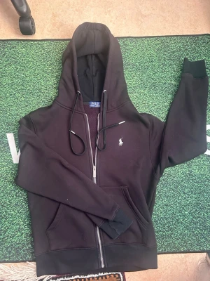 Svart hoodie från Polo Ralph Lauren - Säljer en svart hoodie med dragkedja från Polo Ralph Lauren. Klassisk design med huva, dragsnören och fickor framtill. Ikoniska vita loggan broderad på bröstet. Mjuk insida och ribbade muddar. 