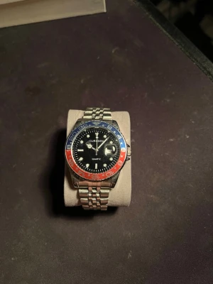 Klocka - Stilren klocka från  med svart urtavla och tvåfärgad vridring i blått och rött. Klockan har ett klassiskt silverfärgat länkarmband i metall och tydliga siffror på vridringen. Fungerar felfritt