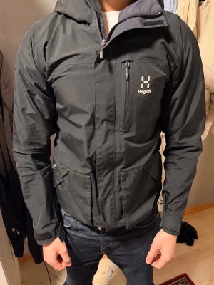 Svart vindjacka från Haglöfs - Snygg svart vindjacka från Haglöfs med huva och dragkedja. Jackan har bröstficka med dragkedja, två sidofickor och justerbara muddar. Perfekt för blåsiga dagar och outdoor-aktiviteter. Tillverkad i slitstarkt syntetmaterial.