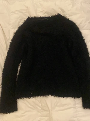 Svart fluffig stickad tröja - Mysig svart stickad tröja med fluffig textur och rund hals. Tröjan har lång ärm och en enkel, rak passform som ger en avslappnad look. Perfekt för kalla dagar när du vill vara både varm och snygg.