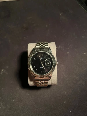 Silverfärgad Seiko herrklocka - Stilren herrklocka med svart urtavla och datumvisning. Klockan har en klassisk länk i silverfärgat metall och markerad räfflad boettkant. Passar perfekt för dig som gillar tidlös design och vill ha en snygg accessoar på handleden. Fungerar felfritt